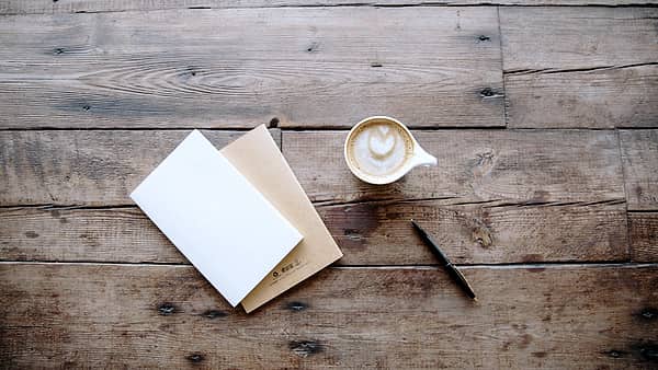 Two notebooks and a pen arranged on a table beside a cup of coffee in daylight