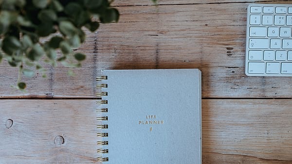 A paper planner lying open on a desk beside a pen, its weekly spread still unmarked