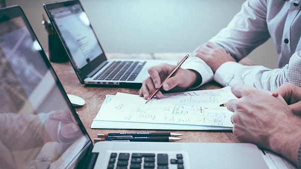 Two people at a table with laptops, one of them marking a printed sheet with a pen