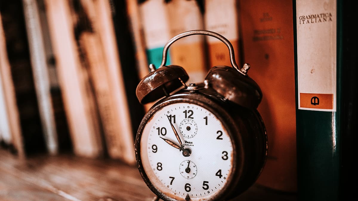 An old mechanical alarm clock standing on a bookshelf beside a row of book spines